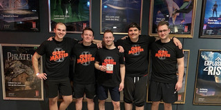 Group of 5 people wearing black T-shirts in front of a wall covered with posters
