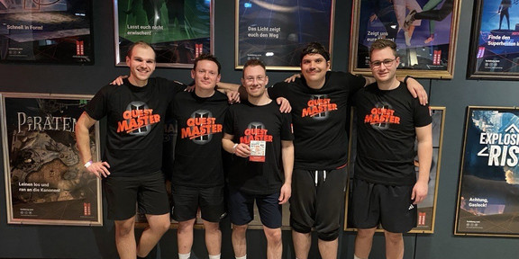Group of 5 people wearing black T-shirts in front of a wall covered with posters